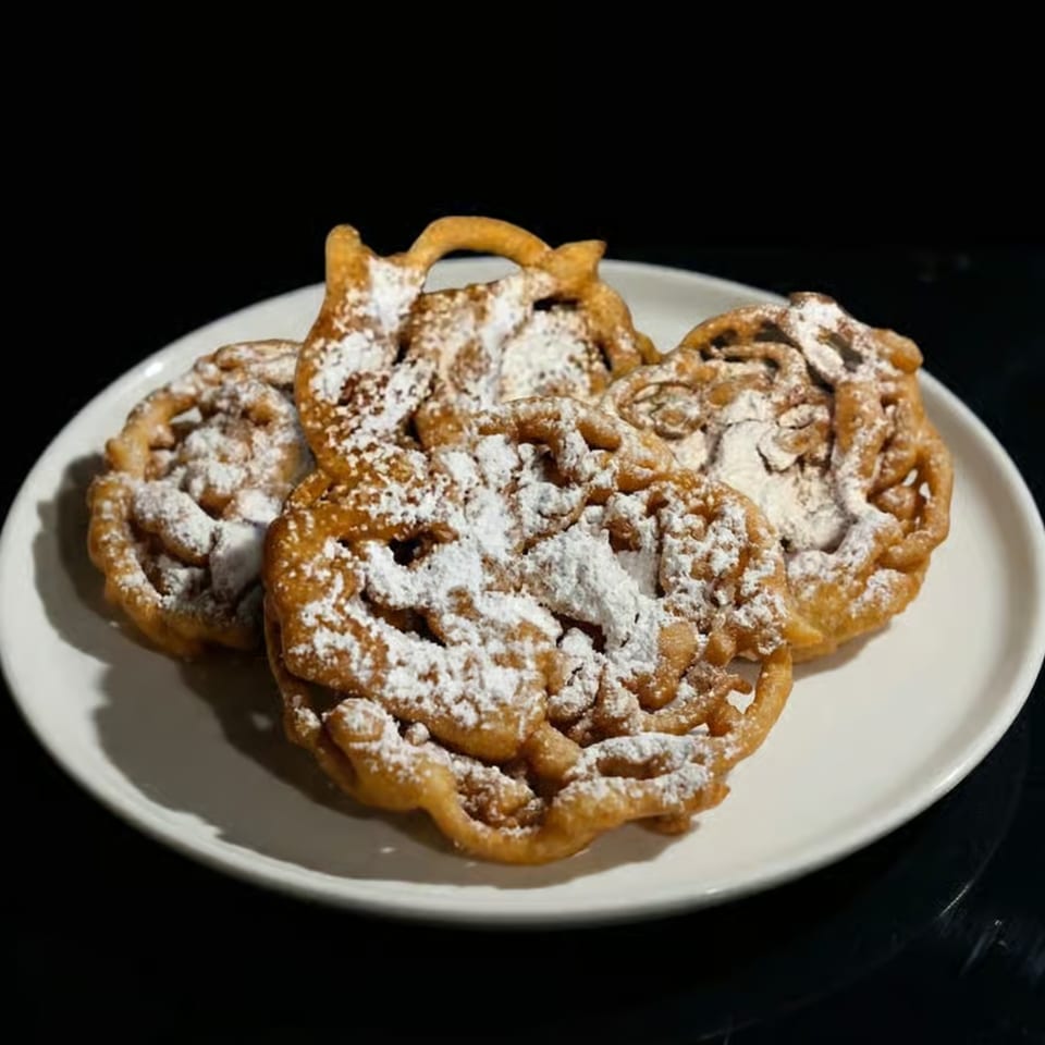 Four golden-brown funnel cakes, dusted with powdered sugar, arranged on a white plate against a dark background.