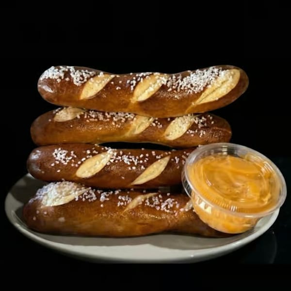 Four salted pretzel sticks stacked on a white plate with a side of cheese dip.