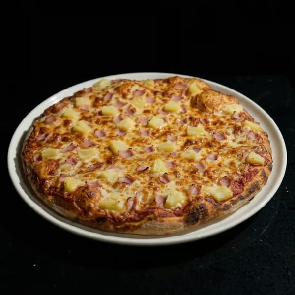 A Hawaiian pizza with ham and pineapple on a white plate against a dark background.