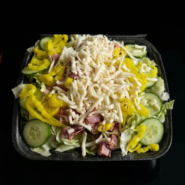 A large salad with lettuce, ham, cucumbers, yellow peppers, and shredded cheese in a black container.